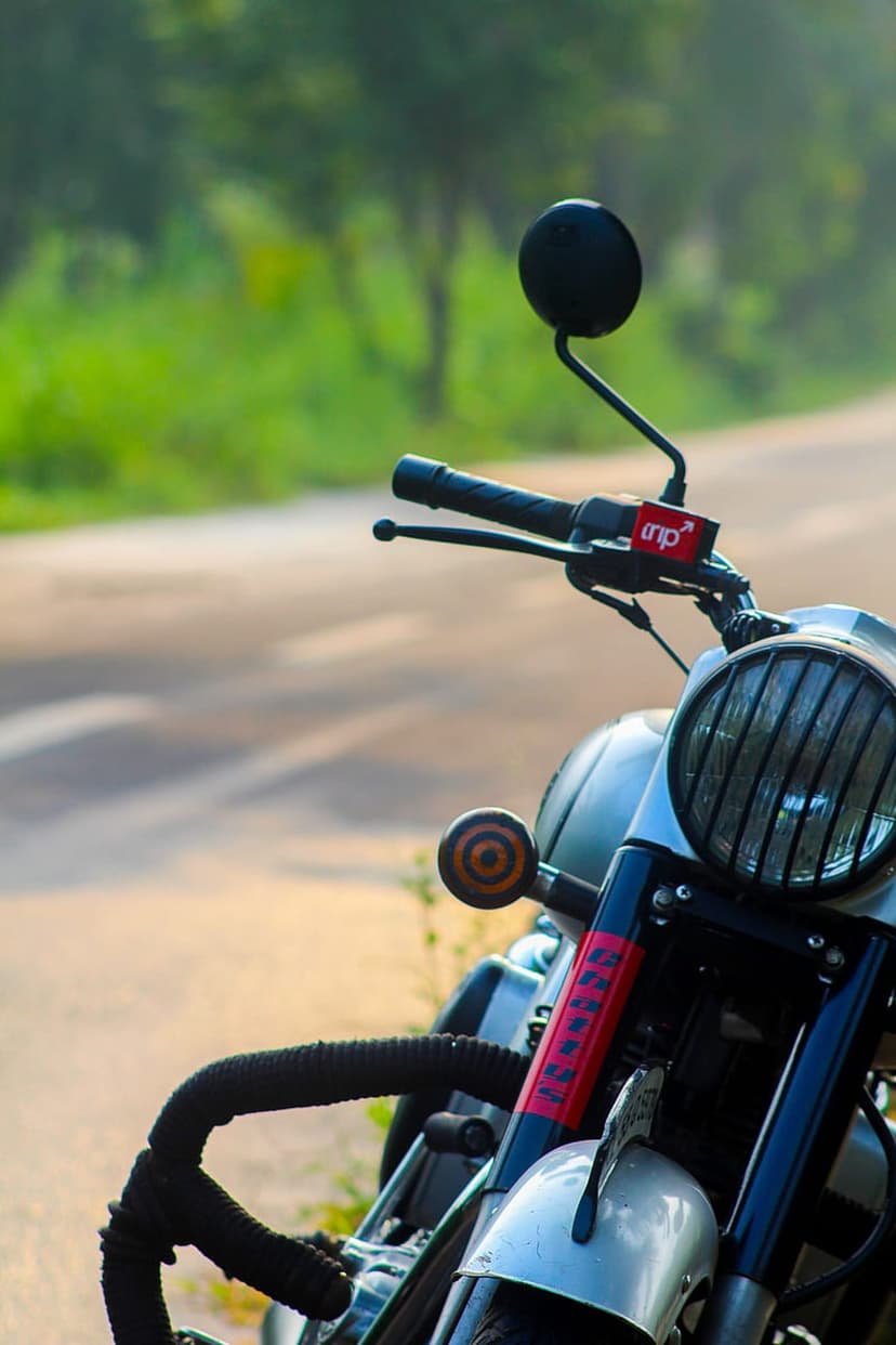 Royal Enfield motorcycle headlight with a black grill and handlebars against a blurred forest road background.