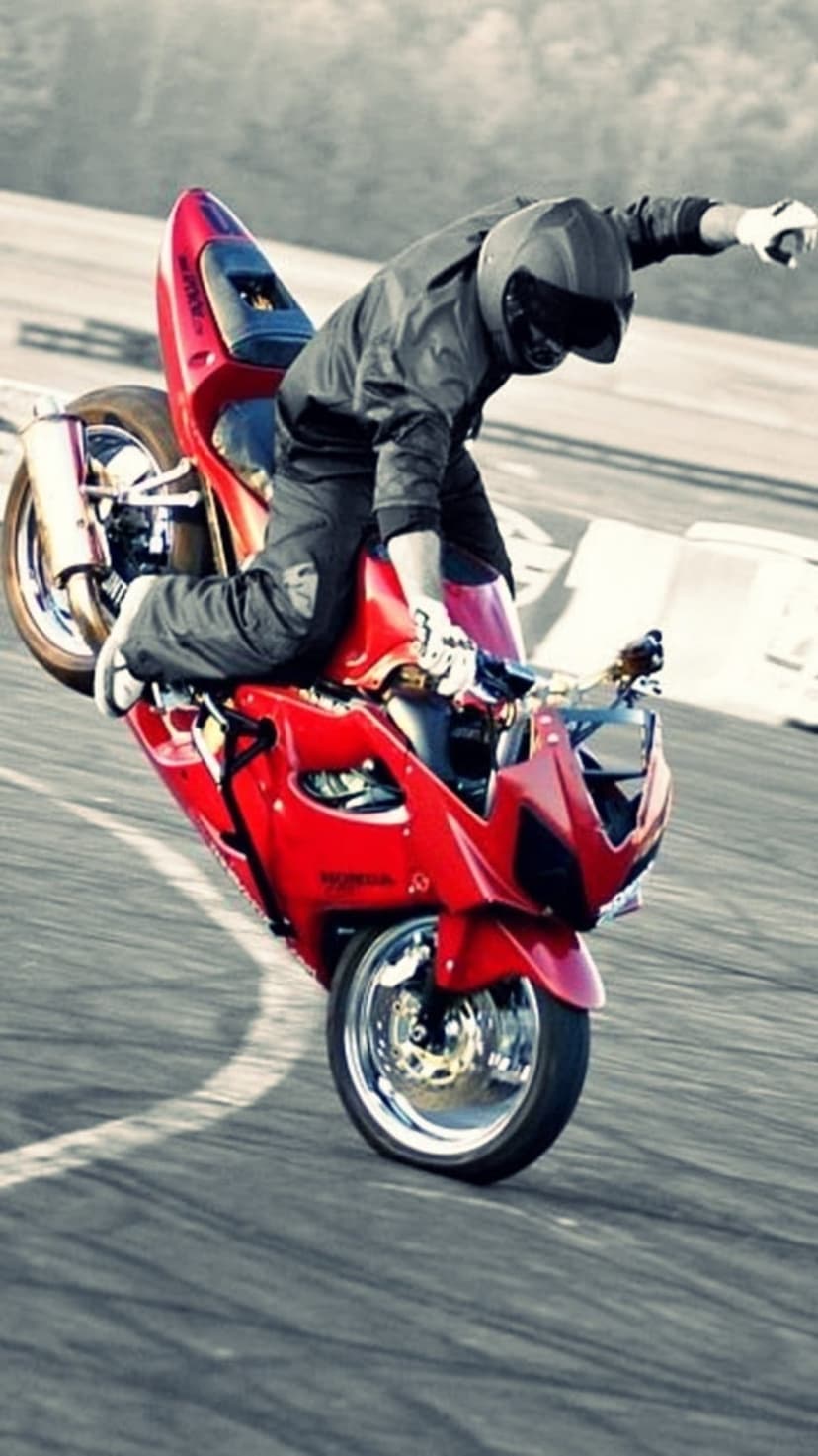Motorcyclist in black gear balances a red sportbike on its rear wheel during a high-speed stunt on a paved track.