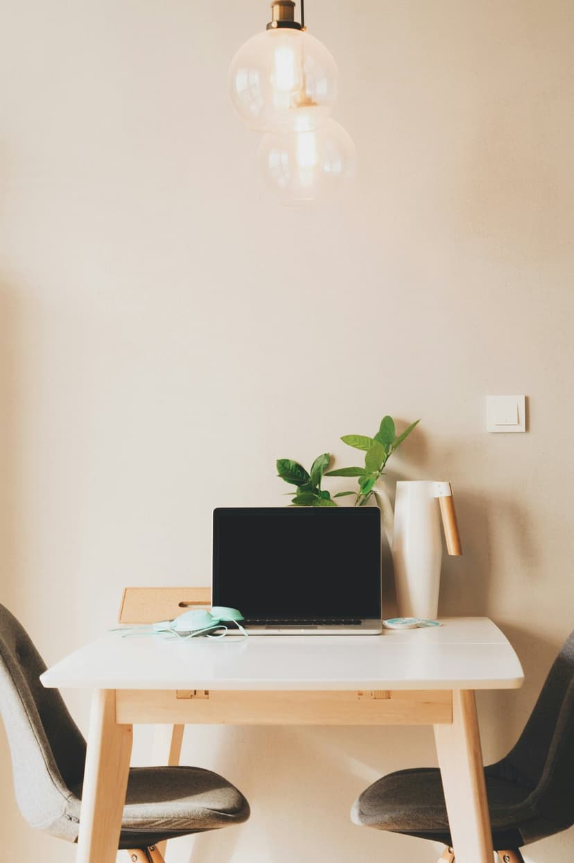 White laptop rests on a modern desk beside a green plant and two grey chairs under a double pendant light.