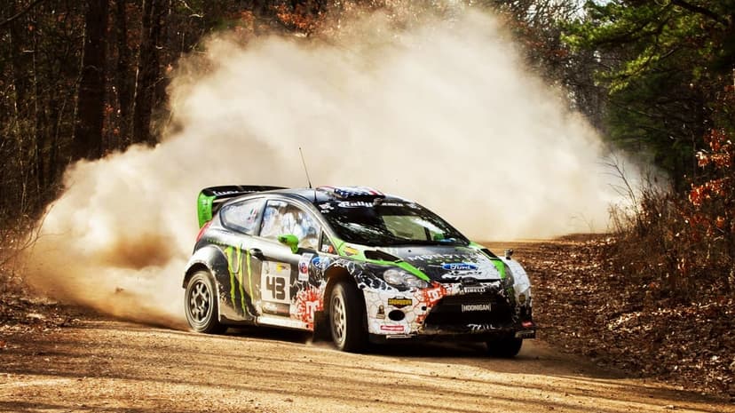 Ford Fiesta rally car 43 drifts on a dirt track, kicking up a massive backlit dust cloud in a dense forest.