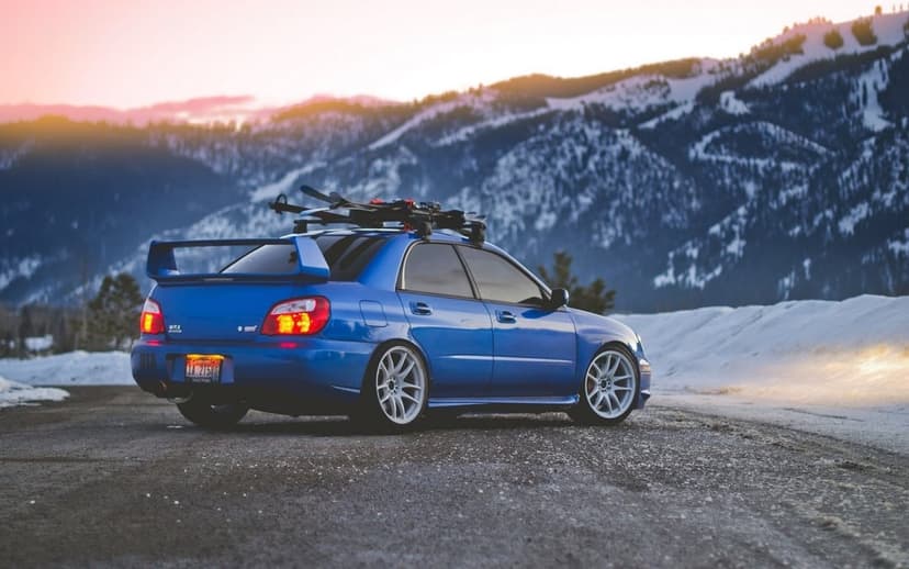 Blue Subaru WRX STi with skis on roof parked on a mountain road at sunset against snow-capped peaks.