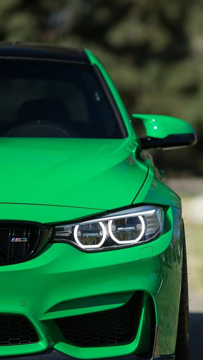 BMW M4 front headlight and green bumper with carbon fiber accents against a blurred natural outdoor background.