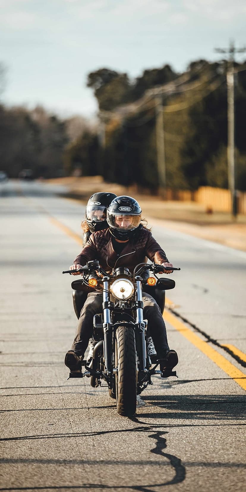 Motorcyclist in a brown jacket and a passenger ride on a paved road with yellow lines under a clear blue sky.