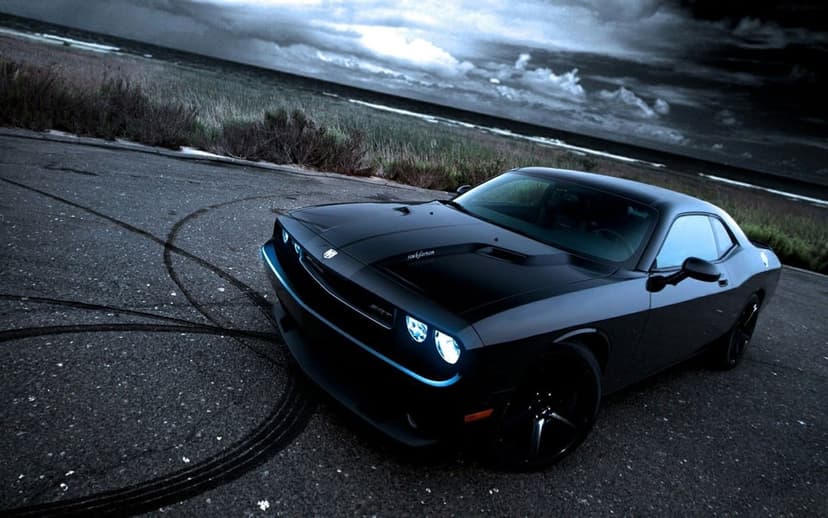 Black Dodge Challenger SRT with glowing headlights on a coastal road under a stormy sky with burnout marks.