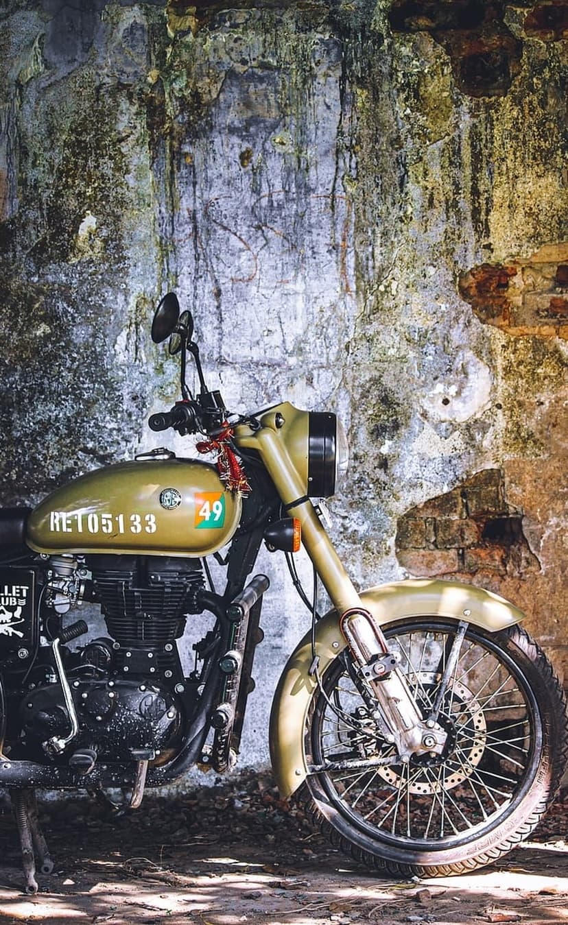 Olive green vintage motorcycle with number 49 parked against a weathered brick wall with moss and peeling paint.