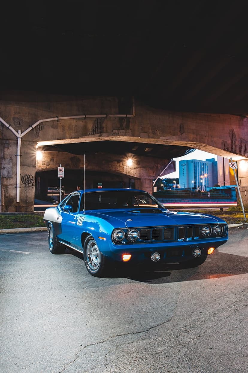 Blue 1970 Plymouth Barracuda parked under a concrete bridge with city lights and chrome accents visible at night.