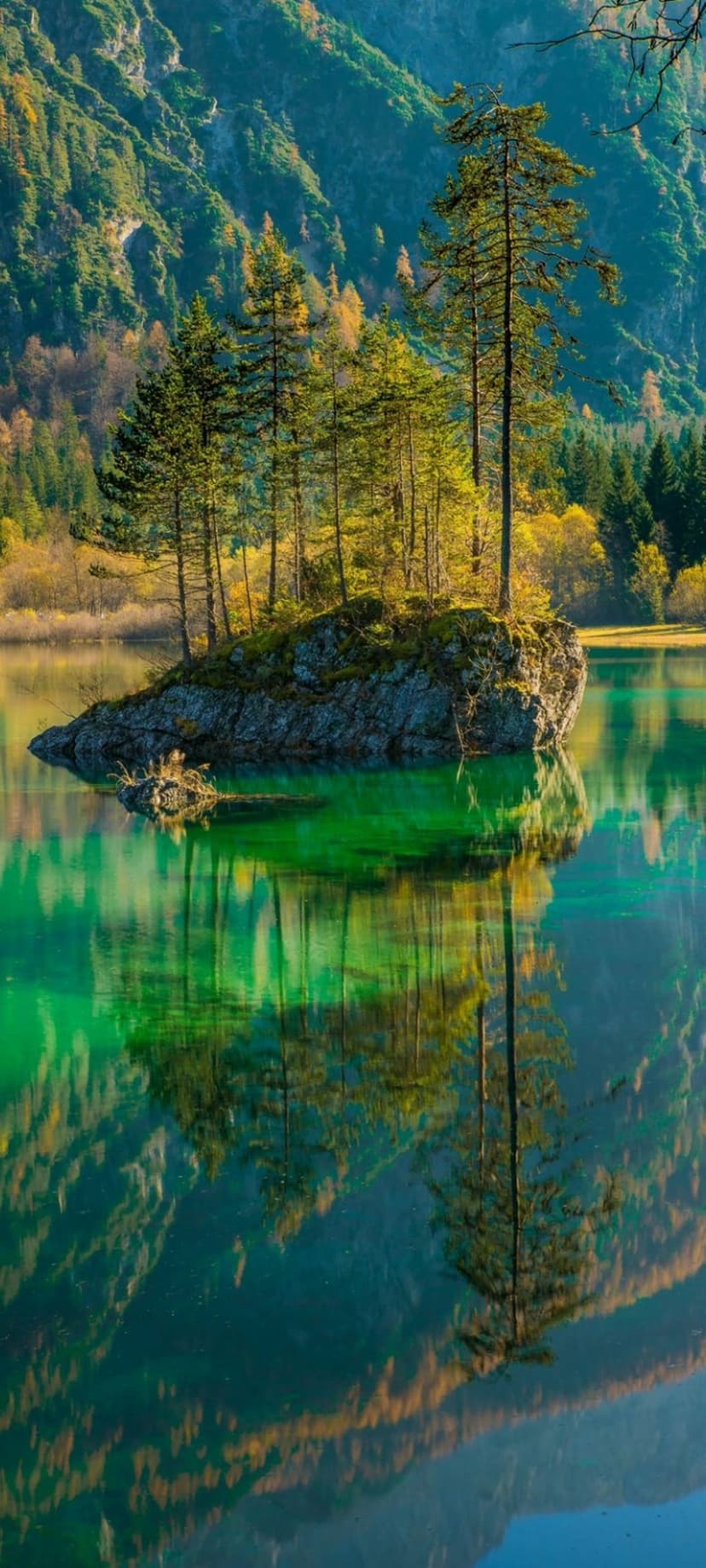 Rocky island with pine trees reflected in clear emerald lake water beneath sunlit mountain slopes.