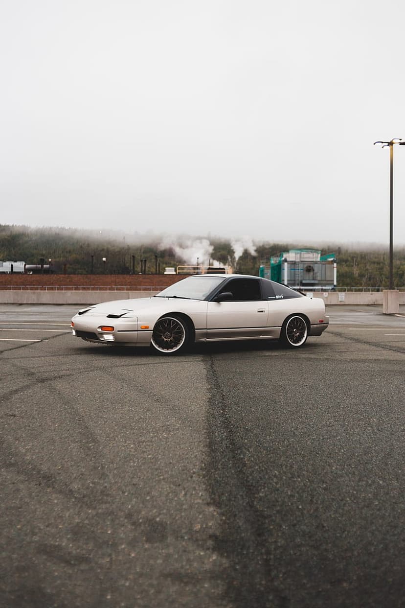 Silver Nissan 240SX with glowing headlights parked on a damp asphalt lot surrounded by thick industrial fog.