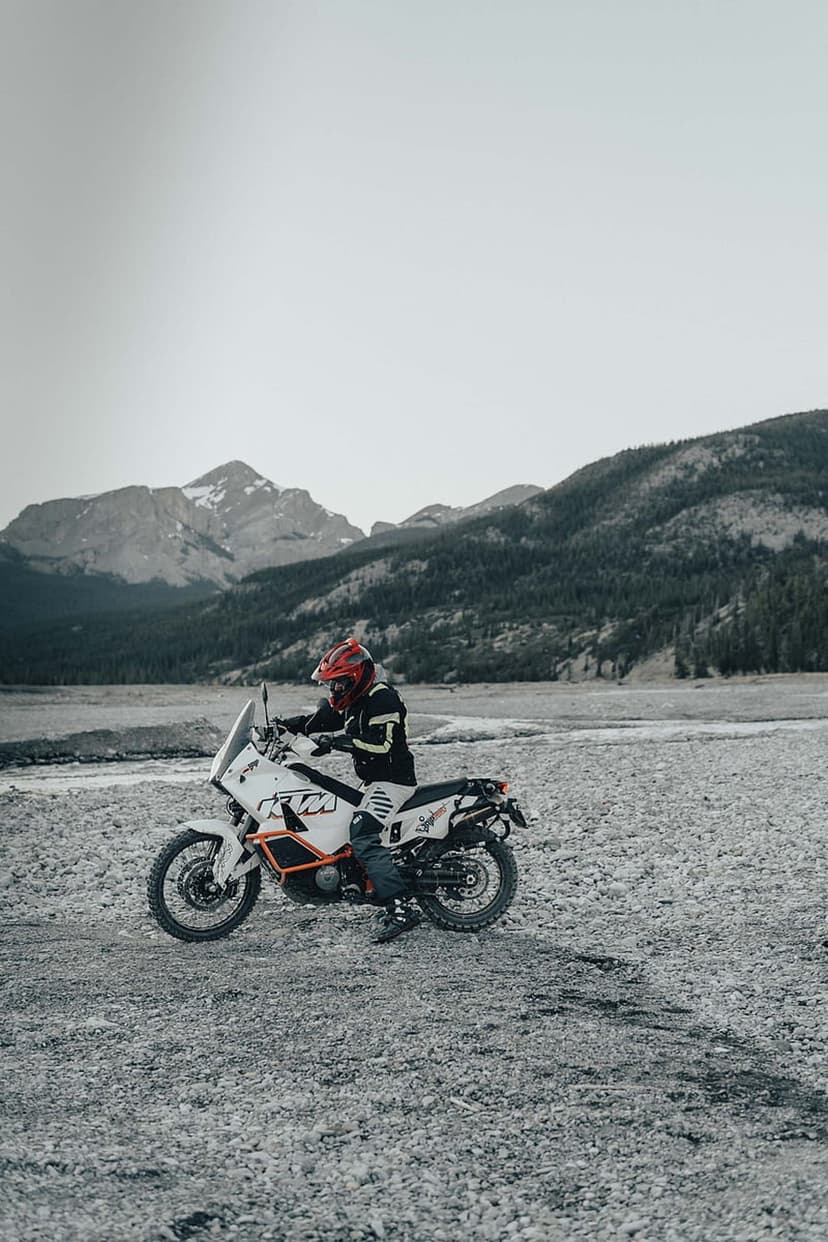 Motorcyclist in a red helmet sits on a white KTM adventure bike within a rocky valley under snowy mountain peaks.