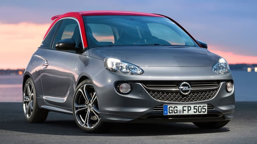 Grey Opel Adam S with a red roof and illuminated headlights parked on asphalt during a coastal sunset.