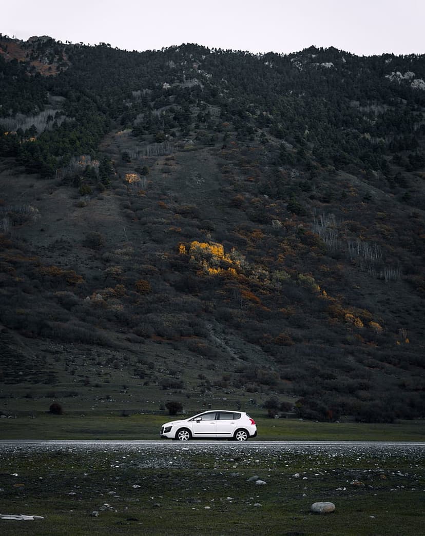 White car on a paved road at the foot of a dark, steep mountain covered in dense trees under an overcast sky.