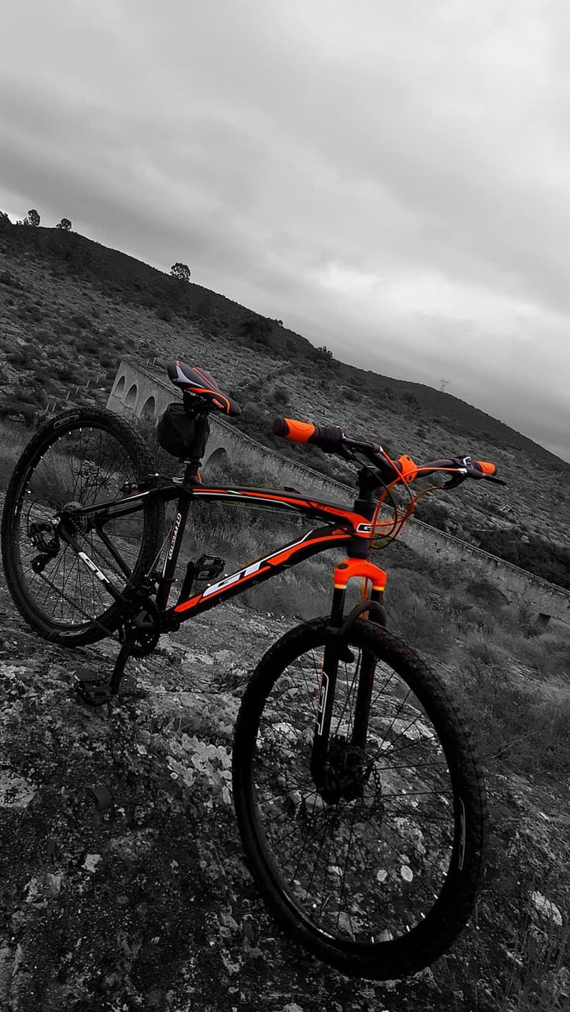 Mountain bike with orange accents rests on a rocky ridge under a cloudy sky near a stone bridge structure.