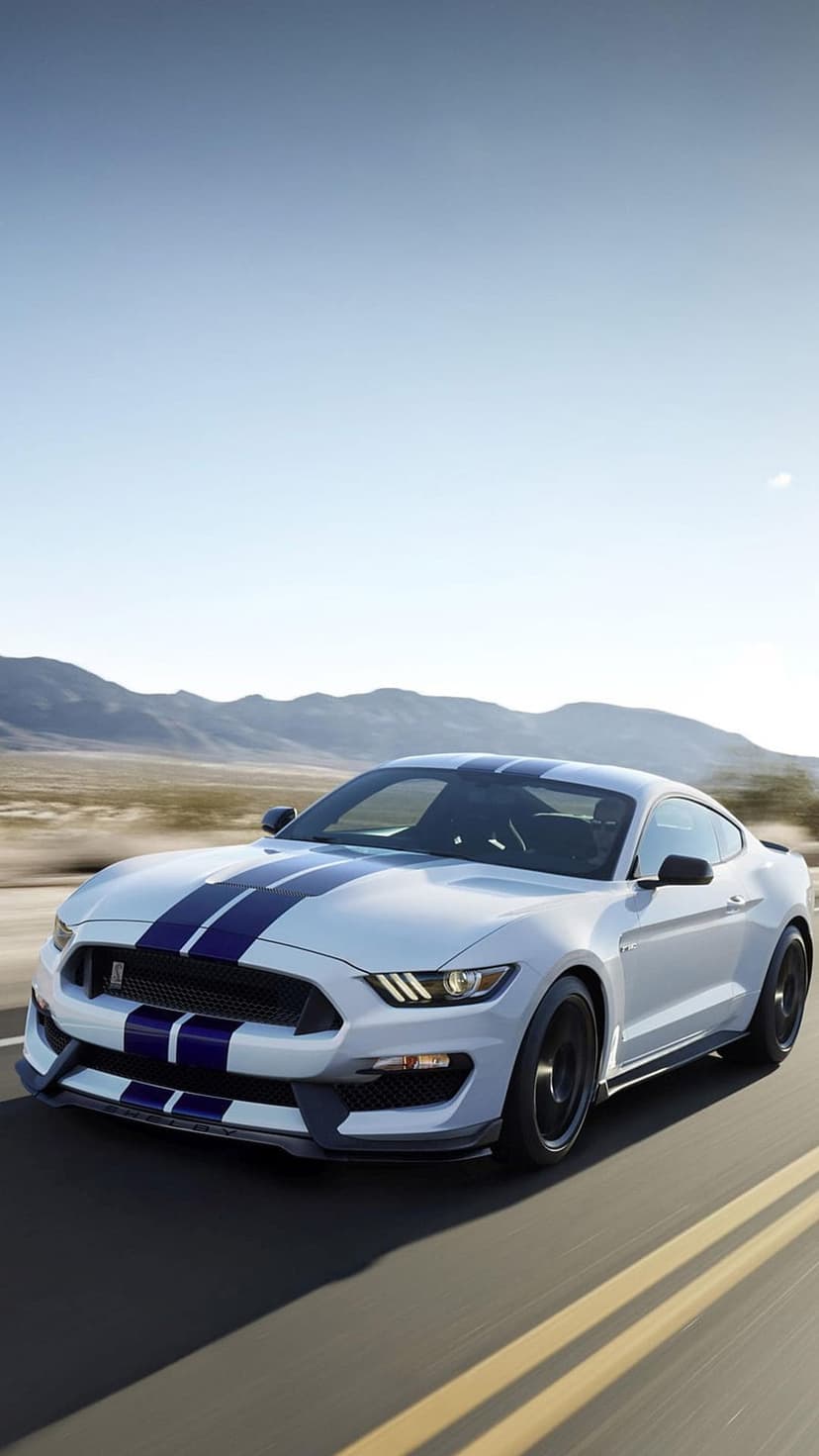 White Ford Mustang GT350 with blue stripes drives on a sunlit desert highway toward distant mountain peaks.