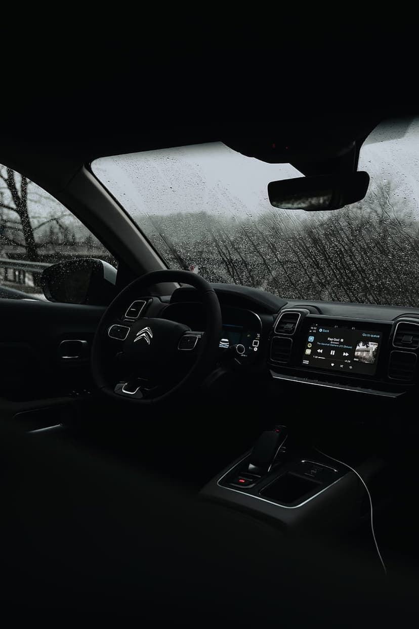 Raindrops on a car windshield frame a dark dashboard and glowing screen with blurred trees in the distance.