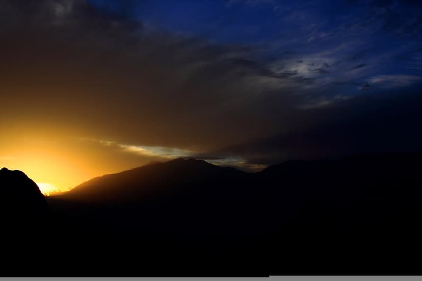 Dark mountain silhouettes stand against a vibrant orange and yellow sunset sky filled with textured clouds.