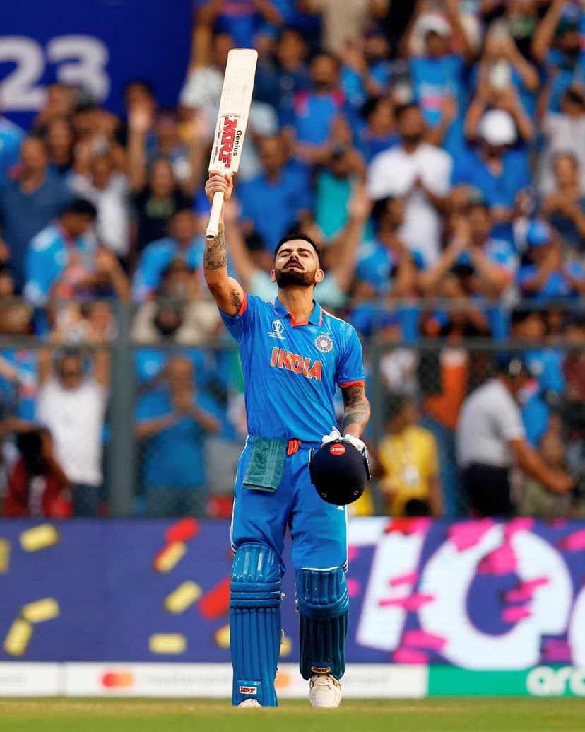 Virat Kohli raises his cricket bat in celebration while wearing a blue India jersey in a crowded stadium.