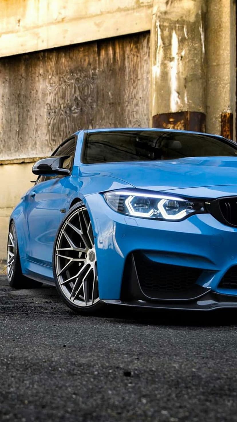 BMW M4 coupe in electric blue with glowing LED headlights parked against a textured gray concrete wall.