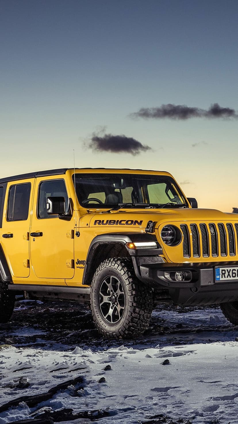 Yellow Jeep Wrangler Rubicon parked on a snow-covered field during a golden sunset with dark clouds.