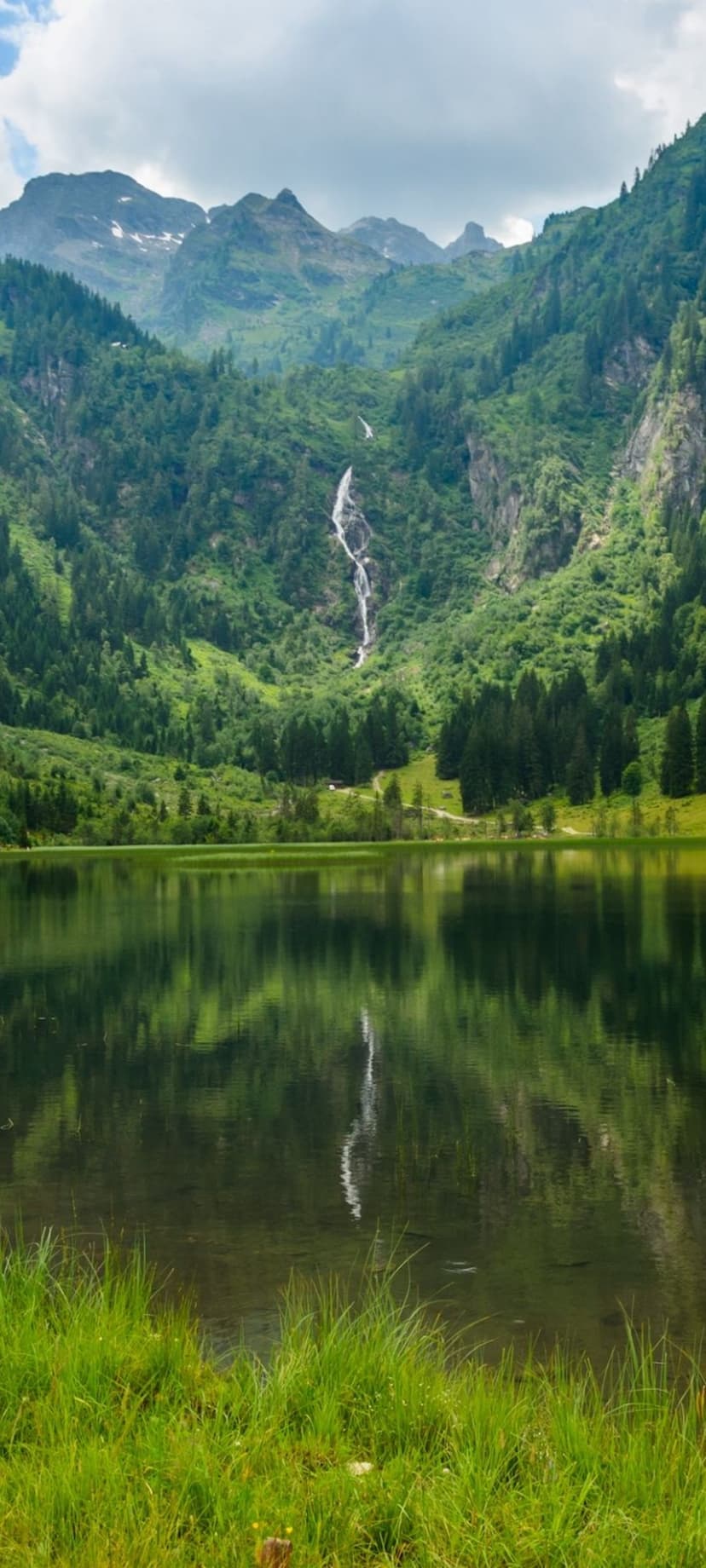 Cascading waterfall flows into a mirror-like lake surrounded by dense evergreen forests under a cloudy sky.