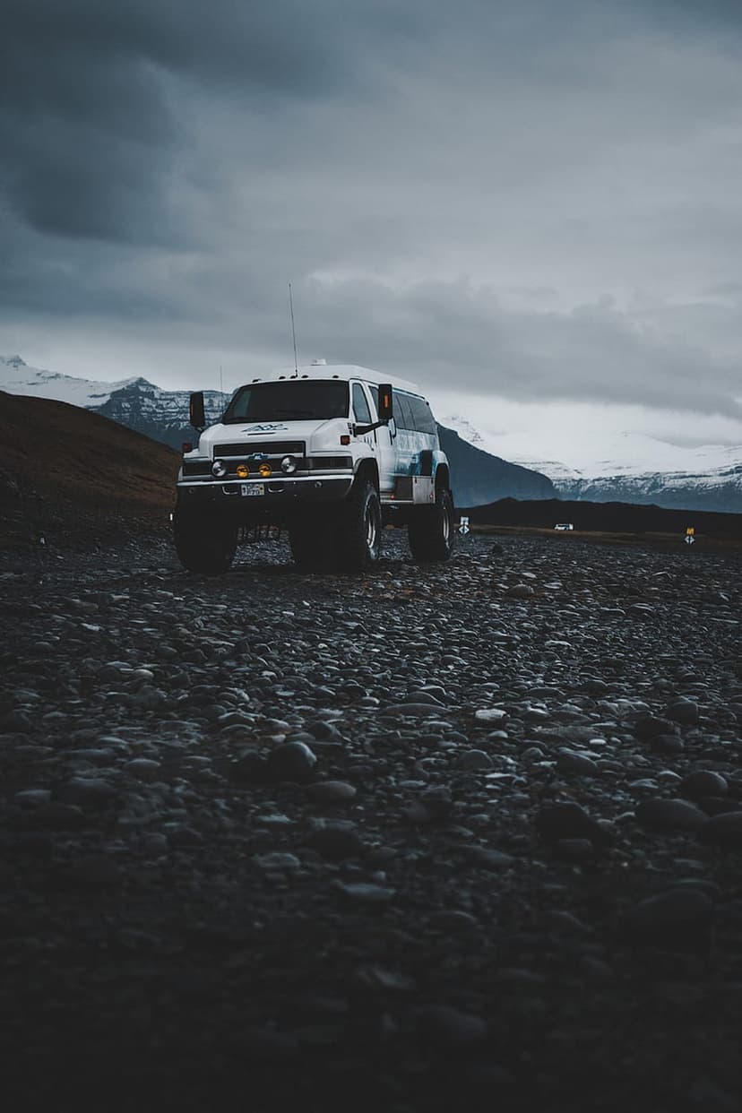 White expedition van with oversized tires parked on dark volcanic rocks before snowy mountains under storm clouds.
