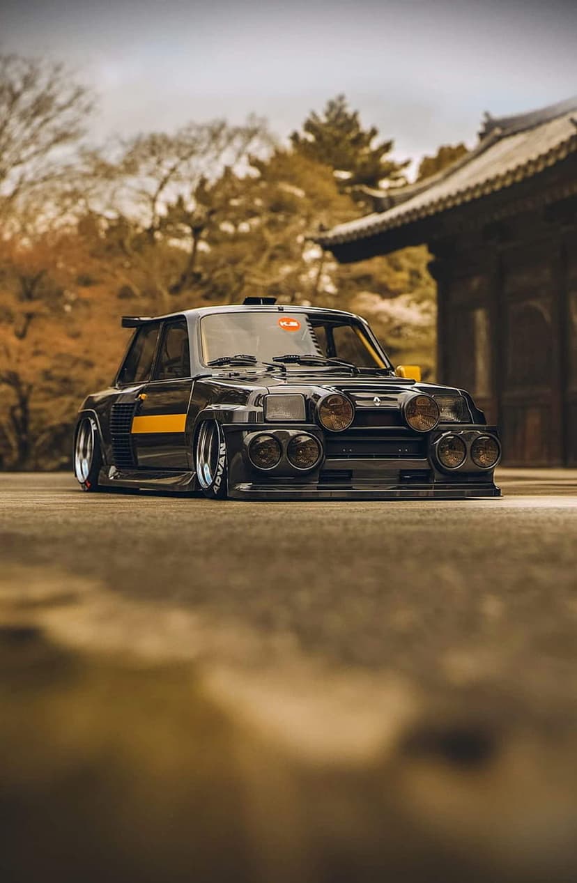 Black Renault 5 Turbo II with yellow stripes parked near a Japanese building among orange autumn leaves.