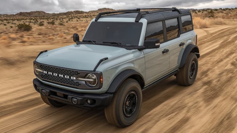 Sage green Ford Bronco driving fast on a sandy desert trail with dust clouds and an overcast sky background.