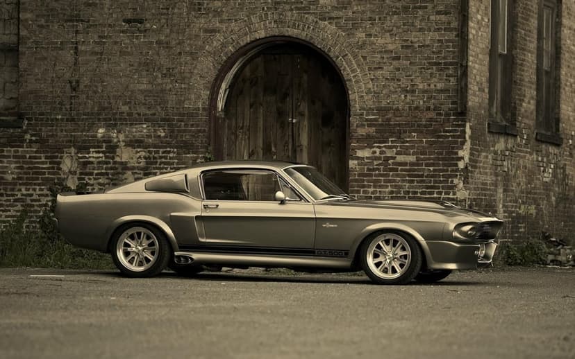 Silver Shelby GT500 Mustang Eleanor parked in front of an old brick building with an arched wooden door.