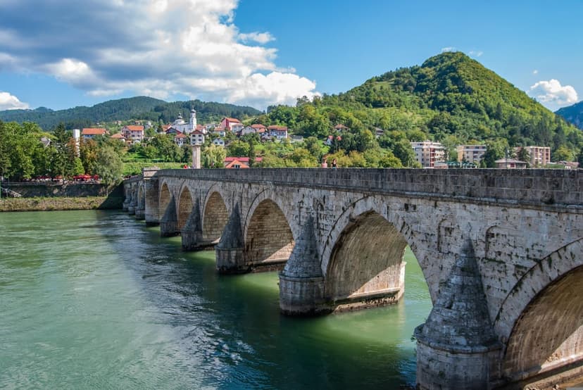 Mehmed Paša Sokolović Bridge with stone arches crossing a turquoise river against green hills in the distance.