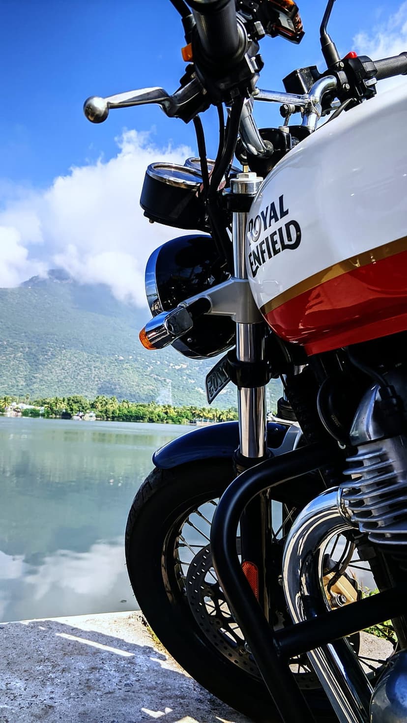 Royal Enfield motorcycle with chrome details parked near a calm lake and green mountains under a bright blue sky.