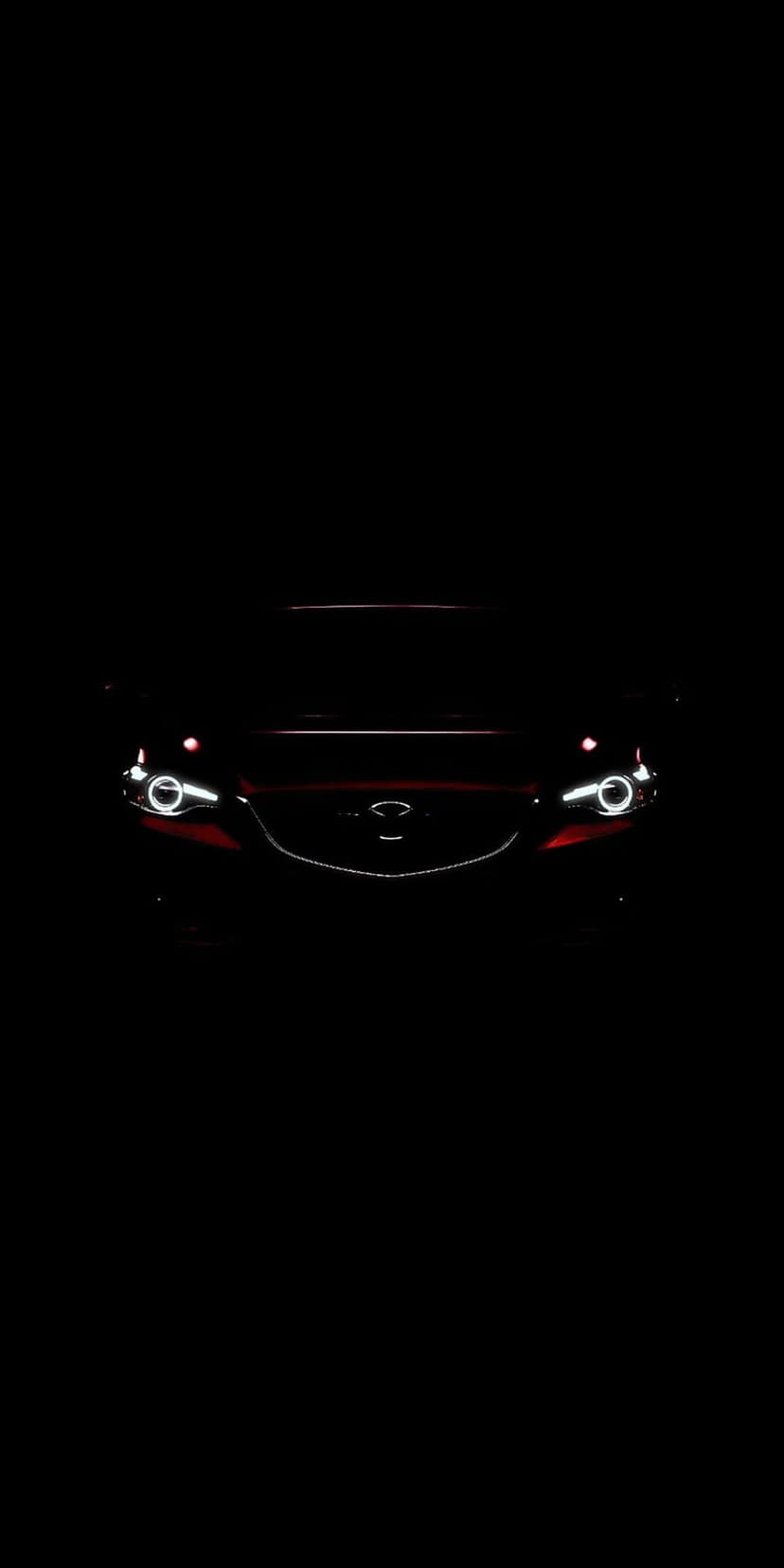Red Mazda front grille and illuminated LED headlights positioned against a dark black background in portrait view.