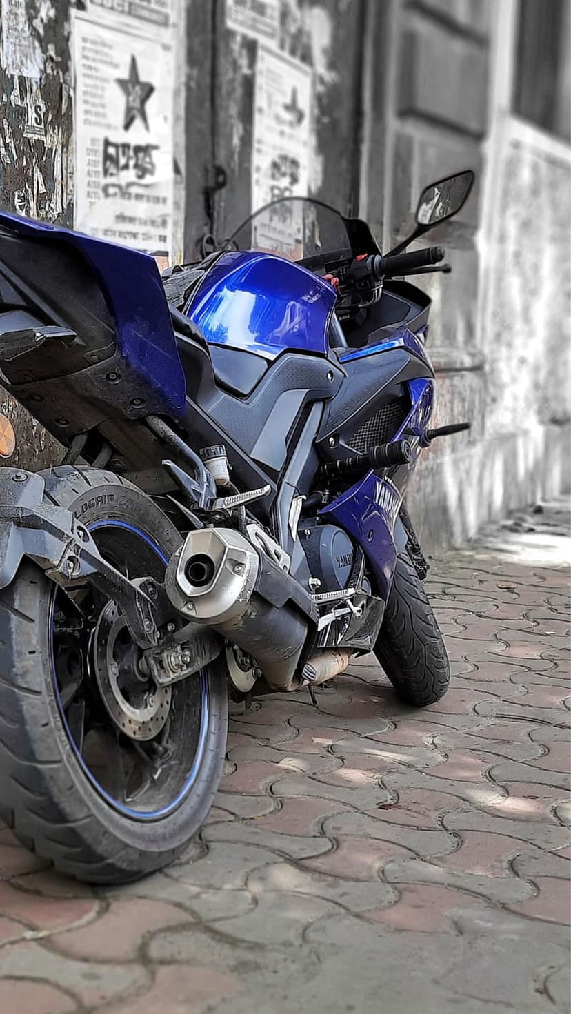 Blue Yamaha R15 motorcycle parked on a brick sidewalk with a visible exhaust pipe and a weathered wall.