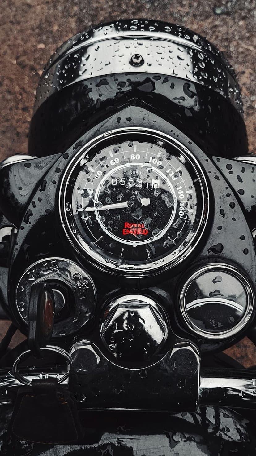 Royal Enfield speedometer with red illuminated logo and chrome accents covered in fresh raindrops after rainfall.