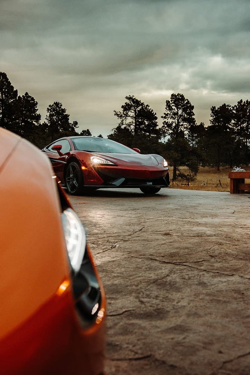 Red McLaren supercar parked on concrete with glowing headlights under a stormy sky and blurred orange foreground.