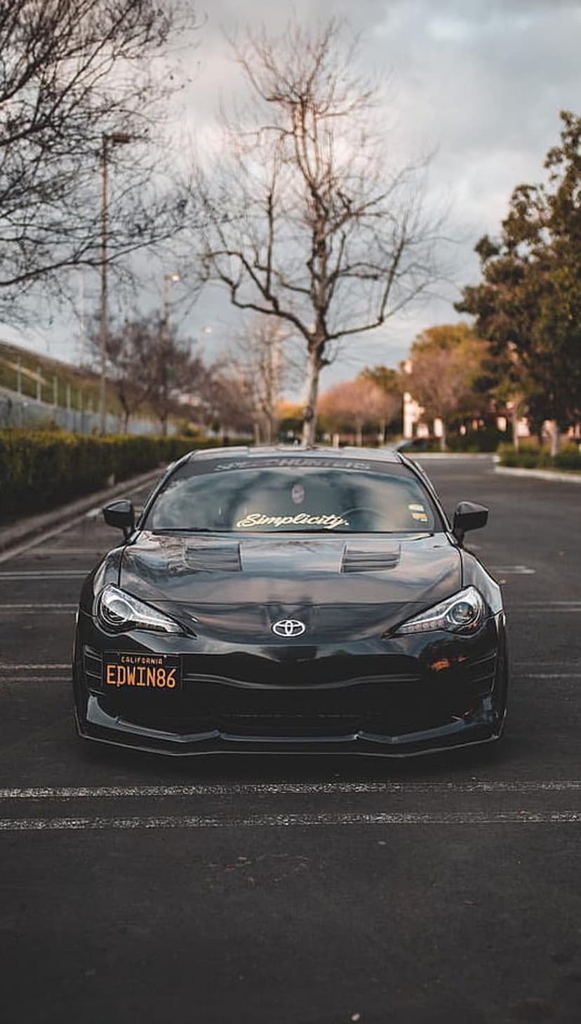 Black Toyota GT86 with a gold Simplicity banner and California license plate parked under a cloudy winter sky.