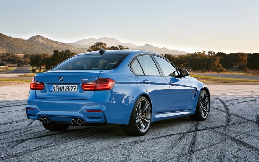 Blue BMW M3 sports sedan rear view on a racetrack with tire marks under a clear sky and distant hills.
