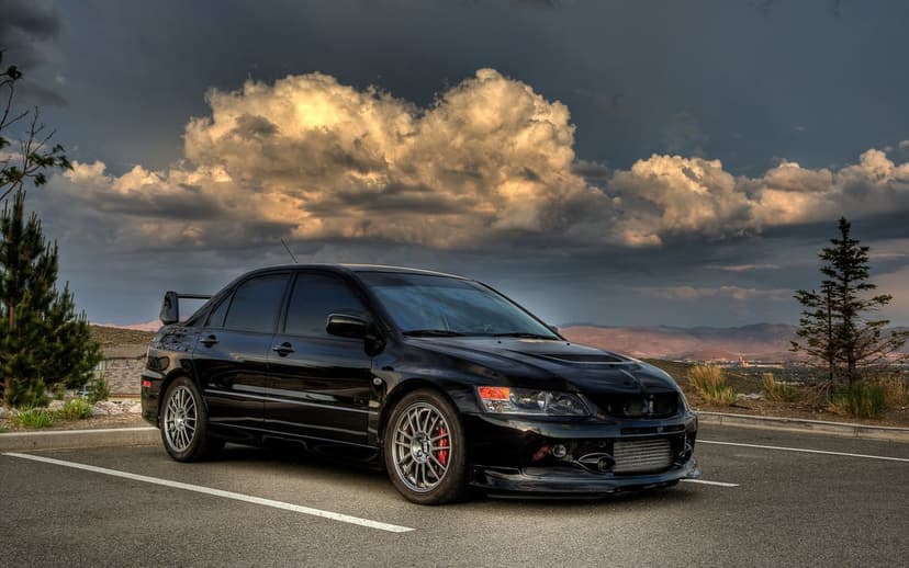 Black Mitsubishi Evolution IX with silver wheels parked on asphalt under dramatic orange and grey storm clouds.