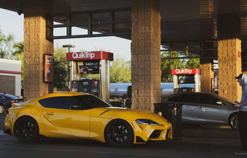 Yellow Toyota Supra sports car parked under a gas station canopy with warm sunlight casting long shadows on asphalt.