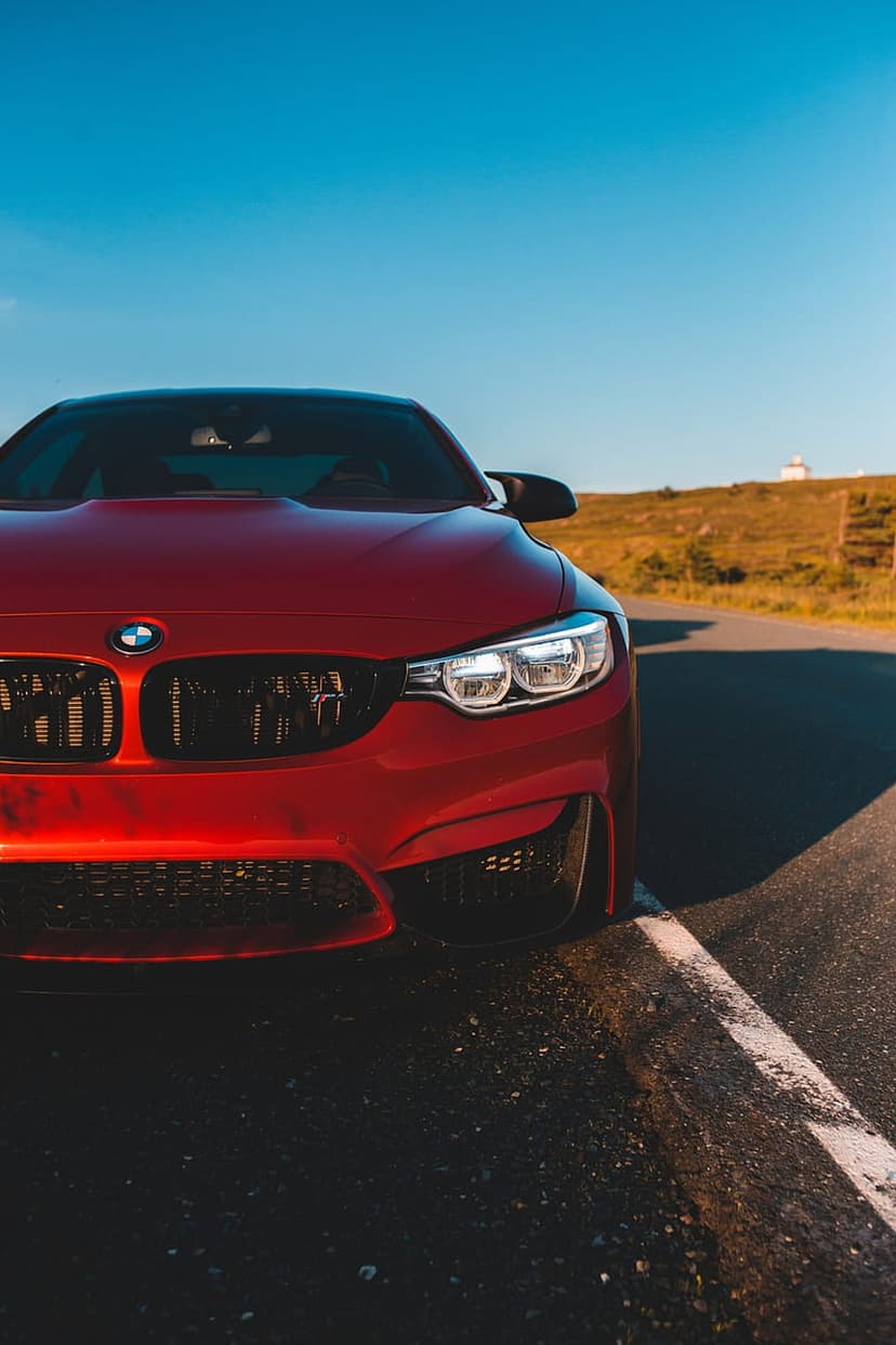 Red BMW M4 sports car on a coastal road at sunset with a lighthouse on a grassy hill in the distant background.