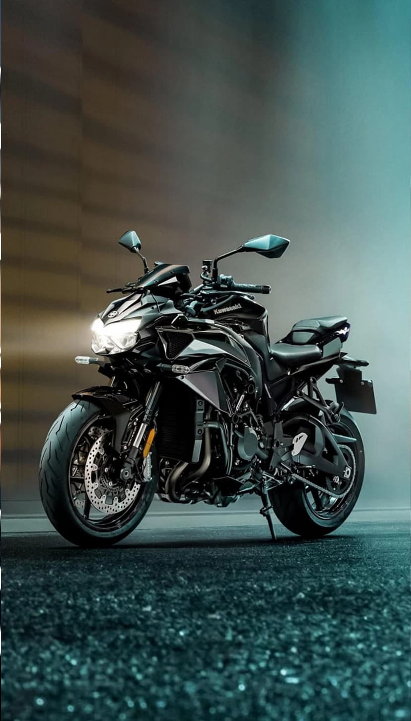 Black Kawasaki motorcycle with a lit headlight stands on a speckled dark floor against a teal and brown background.