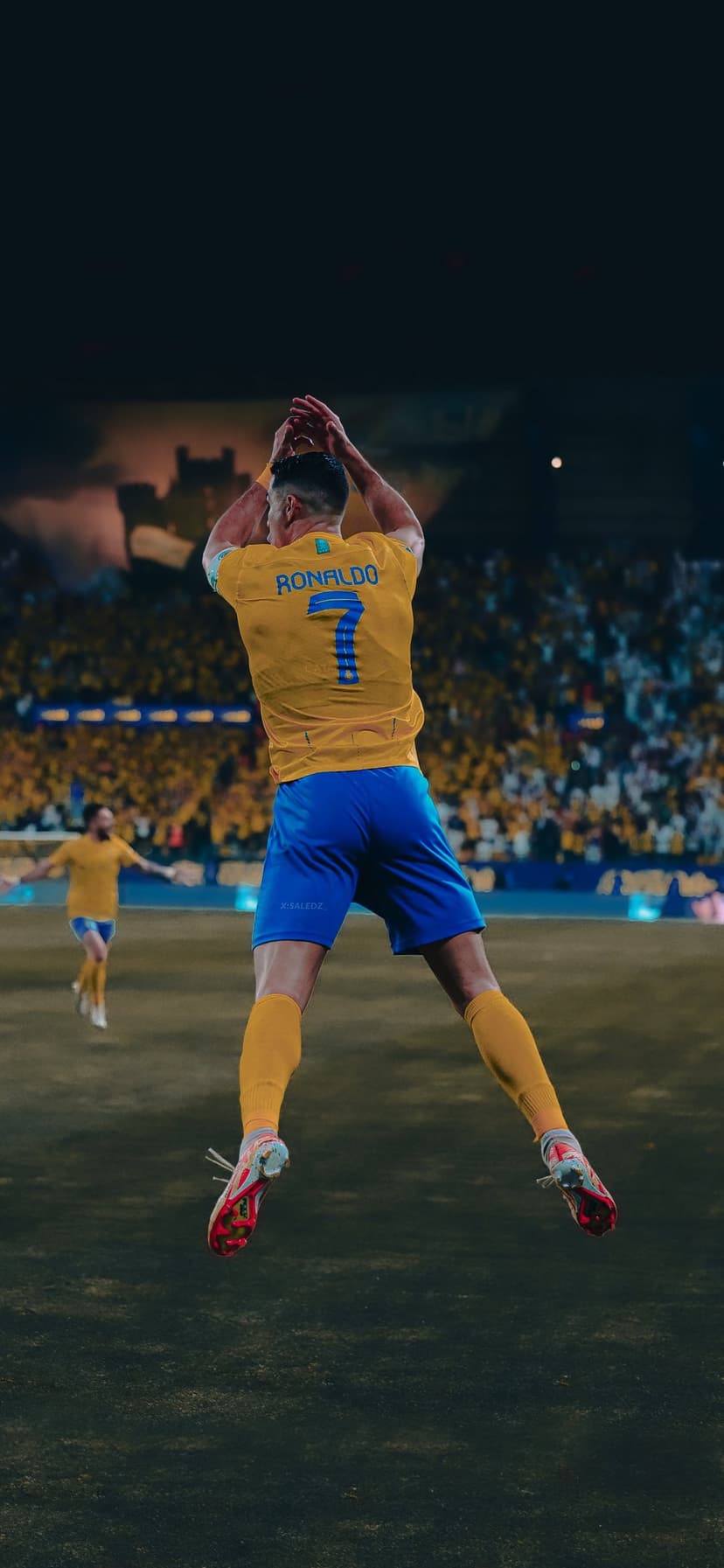 Cristiano Ronaldo jumps mid-air in a yellow and blue kit before a blurred stadium crowd during a match celebration.