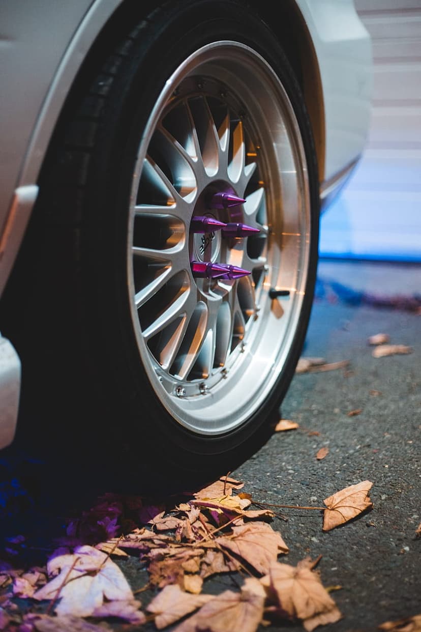 Silver multi-spoke car wheel with purple spiked lug nuts resting on asphalt covered in scattered orange leaves.
