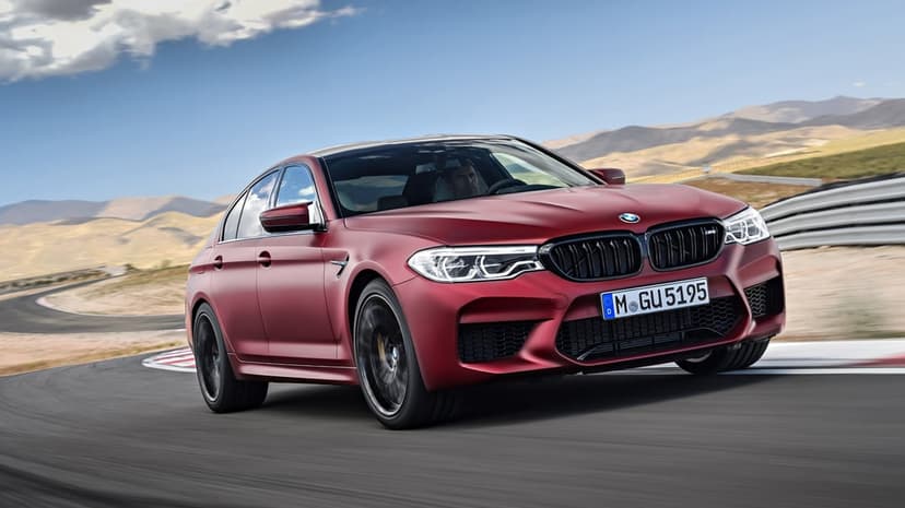 Matte red BMW M5 sedan driving fast on a curved racetrack with mountains and blue sky in the background.