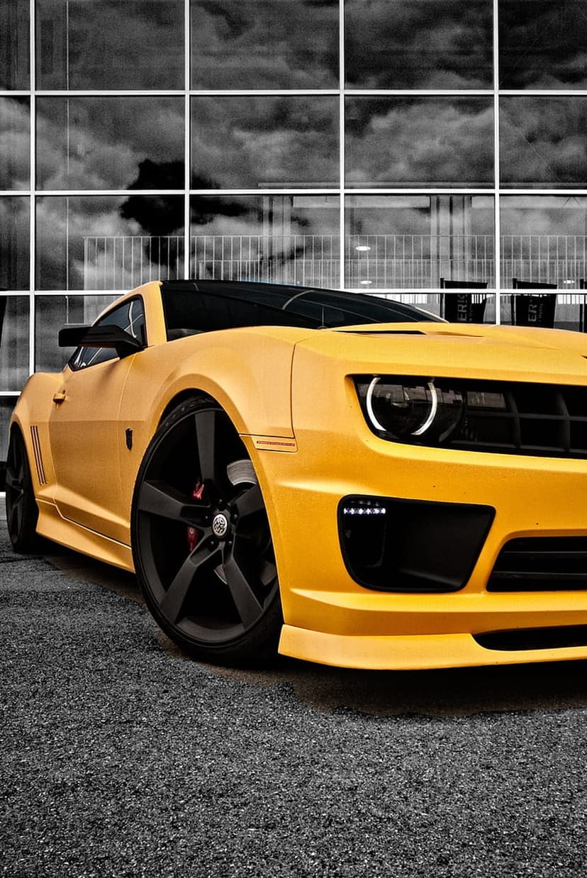 Yellow Chevrolet Camaro with black wheels parked near a grid-glass building under a cloudy sky in a low angle shot