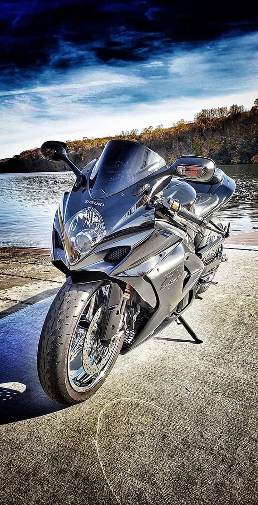 Suzuki sportbike parked on concrete by a calm lake under a dramatic cloudy sky with sunlight breaking through.