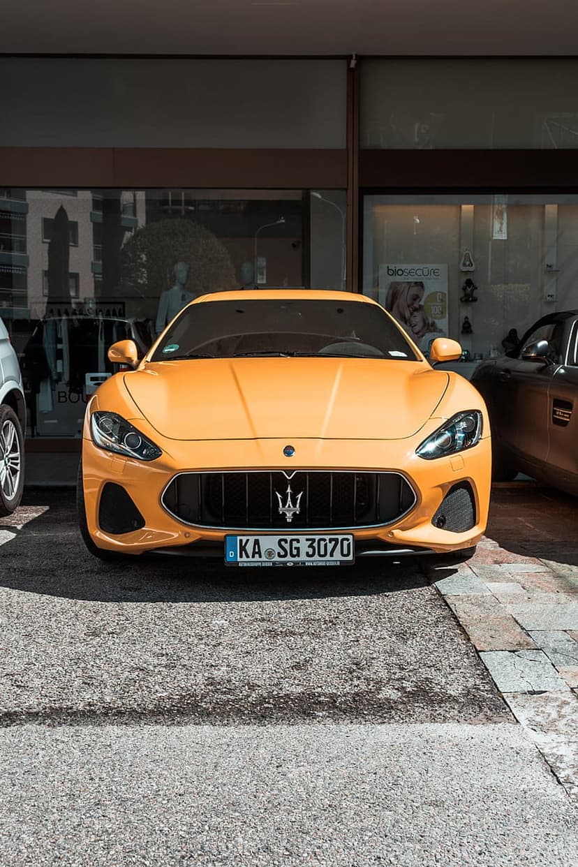Orange Maserati GranTurismo supercar parked on stone pavement in front of a modern glass building storefront.