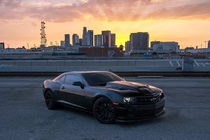 Black Chevrolet Camaro parked on asphalt with the vibrant Los Angeles skyline and sunset in the background.