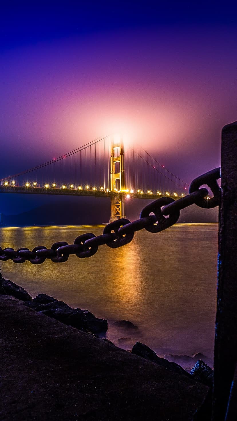 Golden Gate Bridge glows with orange lights behind a thick evening mist and a heavy foreground chain at night.