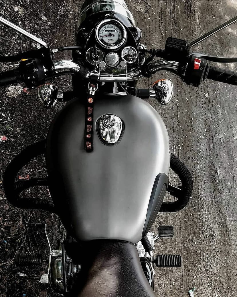 Grey motorcycle fuel tank and chrome handlebars featuring a round analog speedometer and leather keychain.