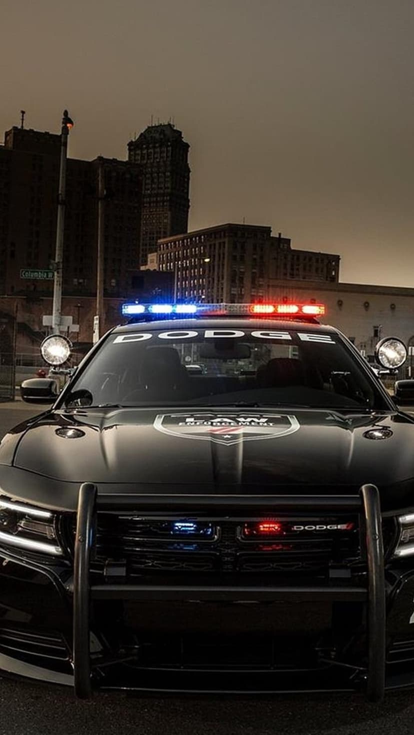 Dodge Charger police car with flashing emergency lights parked before a silhouetted city skyline during twilight.