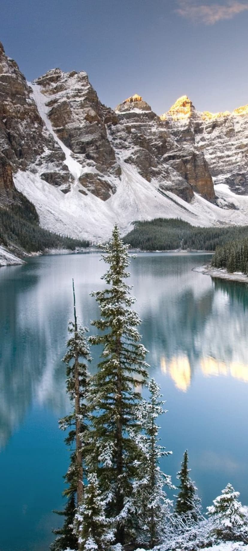 Snow-capped mountain peaks reflect in a turquoise lake with frosted pine trees framing the cold winter landscape.